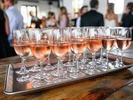 Glasses of pink beverage arranged neatly on a metal tray, set upon a wooden table. The background is softly blurred, revealing people dressed formally in a lively gathering photo
