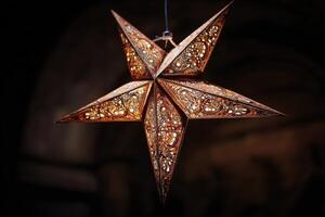 Ornate five-pointed star lantern with intricate cut-out patterns hangs against a dark blurred background, illuminating with a warm glow and creating a cozy ambient atmosphere photo