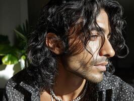 Curly-haired person with long dark hair, wearing a patterned shirt and chain necklace, gazing into sunlight from the side, plant with large leaves slightly blurred in the background photo