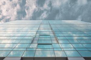 Looking up at a modern glass building reflecting the cloudy sky, creating a seamless illusion of architecture blending with nature. The perspective emphasizes height and transparency photo