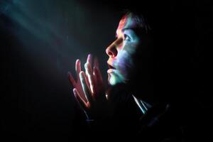 A person gazes upward, hands near face, illuminated by colorful light rays in a dark setting. The light casts vibrant patterns across their face, creating a serene and mysterious mood photo