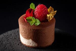 Chocolate mousse cake garnished with a vibrant raspberry, red petals, mint leaves, and gold flakes on a textured surface. The dessert appears rich and elegant, perfect for gourmet presentations photo
