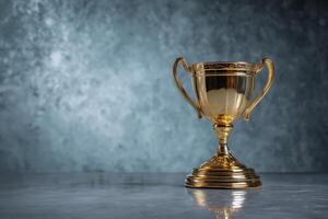 Golden trophy cup with ornate handles and detailed engraving, placed on a reflective marble surface with a smooth, soft-focus background. The cup's shine indicates high value photo