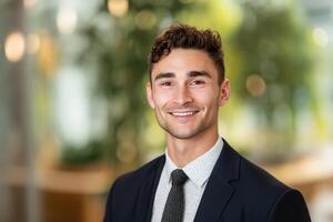 Young man in a business suit smiling confidently. He has short curly hair and is wearing a white shirt with a patterned tie. The setting is indoors with a soft-focus background photo