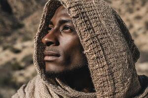 A contemplative person gazes upward, wrapped in a textured, brown hooded shawl. The sunlight casts gentle shadows on their face, highlighting the serene expression against a blurred natural backdrop photo
