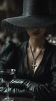 Mysterious woman in black leather outfit, wide-brimmed hat partly hiding her face, wearing a cross necklace with dark lipstick, hands gloved, sitting by a table with a glass of red wine photo