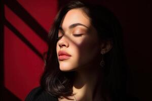 Brunette woman with closed eyes basks in sunlight casting shadow patterns on her face. She wears an earring and dark top. Her serene expression contrasts with vibrant red tones photo
