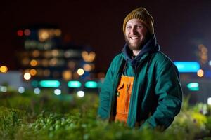 Smiling man in green jacket and orange overalls standing in a lush green garden at night. He wears a knitted hat, city lights softly illuminate the background with a gentle bokeh effect photo