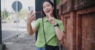 Cheerful young woman happily takes a selfie while walking along a sidewalk next to a brick wall. video