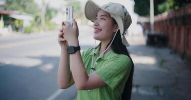 asiático mujer en verde camisa y sombrero en pie en el lado de el calle disfrutando el ciudad ver y tomando fotos con móvil teléfono. video