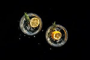 Two glasses present cocktails with lemon slices and sprigs against blackness, droplets around photo