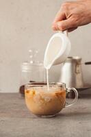 Hand pouring milk into a glass mug with coffee and ice. Cold Brew coffee making process. Vertical background. Copy space for text photo