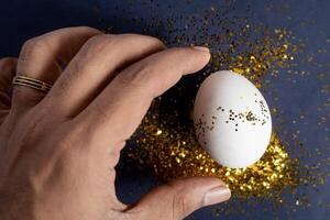 Hand touching an egg decorated with gold glitter on a dark background. photo