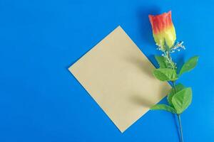 Colorful artificial rose flower and yellow paper blank card on dark blue background. photo