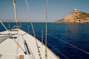 view from a sailing boat to an island with a lighthouse in clear sunny weather, the bow of the boat, the shrouds of the yacht, reflection on the water, mountains in the background photo