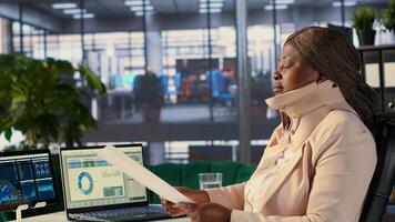 CEO with cervical collar evaluating employees performances based on monthly reports, doing general analysis while she wears the medical neck foam after injury. Director examines trends. Camera A. photo