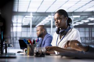 African american engineer next to coworkers in server farm checking recovery plan, monitoring data center energy consumption. Woman in server hub doing routine disk checks photo