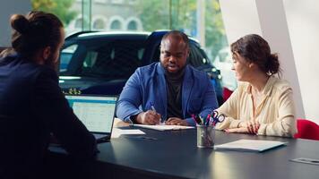 Car dealership salesman sitting with clients at table, giving them contract to sign after finishing test drive. Agent suggesting couple extra vehicle features to buy, waiting for signature, camera A photo