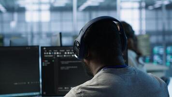 System administrator working in computer network security data center listening music. African american worker wearing headphones monitoring server racks energy consumption, camera A photo