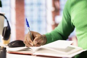 Man reading and writing for school exam on notepad at home office desk, close up. African american student doing homework, writing down info with pen, researching for university photo