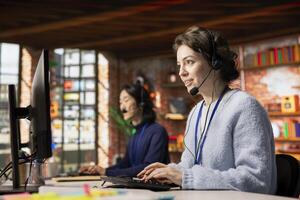 Jolly call center worker dealing with upset customers, speaking using headset at office desk. Friendly woman doing customer support job in workplace using computer, handling clients complaints photo