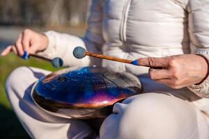 Close-up of hands playing a steel tongue drum with mallets against the backdrop of Altai mountains and river. Concept of sacred sound, touch of nature, and mindful musical ritual. High quality photo