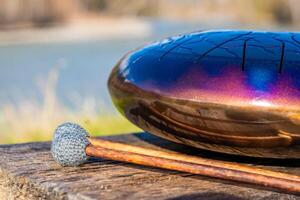 Steel tongue drum and mallets resting on a wooden bench with mountains, river, and forest in the background. Concept of sacred stillness, sound meditation, and harmony with nature. High quality photo
