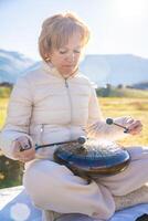 Woman playing a steel tongue drum in nature with a mountain river and Altai landscape in the background. Concept of harmony, mindfulness, and connection with nature. High quality photo