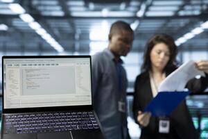 Close up of code lines on laptop screen in front of data center employees reading paperwork. Focus on programming language on notebook display next to server hub workers reading technical files photo