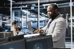 Data center admin using laptop to update servers tech, configuring gear using software. Server farm IT professional using notebook to review hardware analytics, ensuring reliable network operations photo
