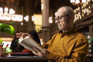 Senior person enjoying lecture at a public library and reading many books, feeling relaxed and increasing his knowledge during retirement period. Happy pensioner with spectacles, hobby. photo