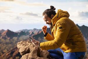 Male hiker reading some good news and feeling joyful with his phone, being distracted by the strong signal and internet connection. Modern explorer cheering with excitement on mountain peak. photo