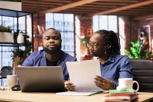 Black remote employees organizing calendar appointments using laptop, checking their papers and synchronizing important tasks on schedule. Man and woman couple review files at home. photo