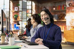 Portrait of happy call center consultant handling customer inquiries, complaints and support requests in office. Cheerful Asian man doing customer service job assisting clients with issues related to products photo