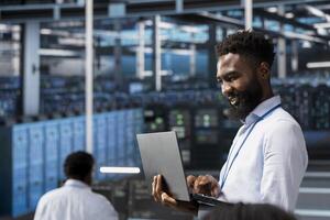 Jolly data center software developer monitoring system performance with laptop, identifying bottlenecks. Joyous server farm IT professional troubleshooting glitches, implementing optimizations photo