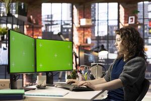Professional woman works at her modern workspace using green screen on dual monitors setup. High tech office environment enhances productivity, ideal for IT professionals and content creation. photo