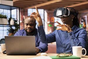 Black girl engages with virtual reality while her partner works on a laptop at the desk, exploring AI and interactive technology in a modern home office setting. 3D simulation with gadgets. photo