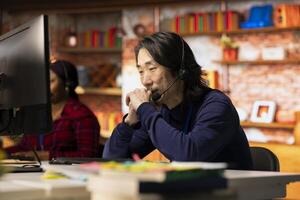 Call center worker dealing with upset customers, speaking with them online using headset at office desk. Asian man using PC to do customer support job in workplace, handling clients complaints photo