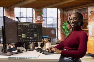 Portrait of upbeat woman working in innovative startup office using programming languages to build application. Jolly African american software development agency IT expert optimizing code on computer photo