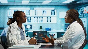 Black general practitioner explaining clinical records and results to a patient during a health care visit, focusing on treatment advice and effective communication in a facility office. Camera B. photo
