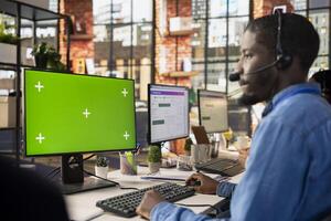 Call center worker in office solving customers issues using green screen PC. African american man in customer support job in front of chroma key computer display using headset to talk with clients photo
