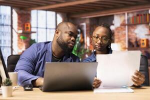 Young people completing spreadsheets on laptop by entering numerical data from papers, calculating totals and analyzing figures. African american couple working together from home. photo