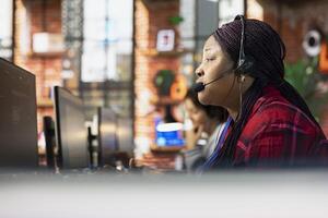 Call center representative in office helping customers by answering questions, close up shot. African american woman in customer support workspace using headset to talk with clients photo