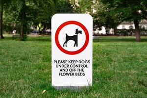 Sign in a park asking to control dogs and keep them off flower beds. Surrounded by green grass trees and public space.12 photo