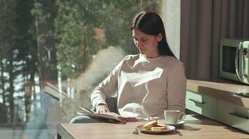 Young woman enjoying peaceful morning, reading book while having breakfast with tea and pastry in sunlit modern kitchen overlooking natural scenery video