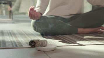 Sunlight illuminates a woman practicing yoga and meditation with her hands in a mudra position, while a smoldering white sage bundle rests on a small plate in front of her video