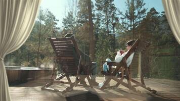 Sunlight streams through curtains onto a wooden deck where a couple relaxes in rocking chairs, enjoying the serene view of a steaming hot tub and a tranquil forest video