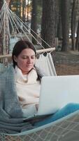 Woman freelancing remotely from cozy hammock, working on laptop while nestled in serene snowy forest landscape, embodying digital nomad lifestyle and work-life balance video