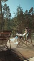 Comfortable woman wearing white boots and a cream-colored sweater enjoys a warm beverage while sitting on a wooden deck chair, taking in the serene view of a tranquil mountain landscape video