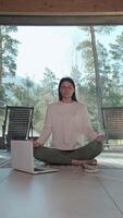 Yoga instructor following online course on laptop, meditating in lotus position with incense burning near her, in front of large windows overlooking mountain landscape video